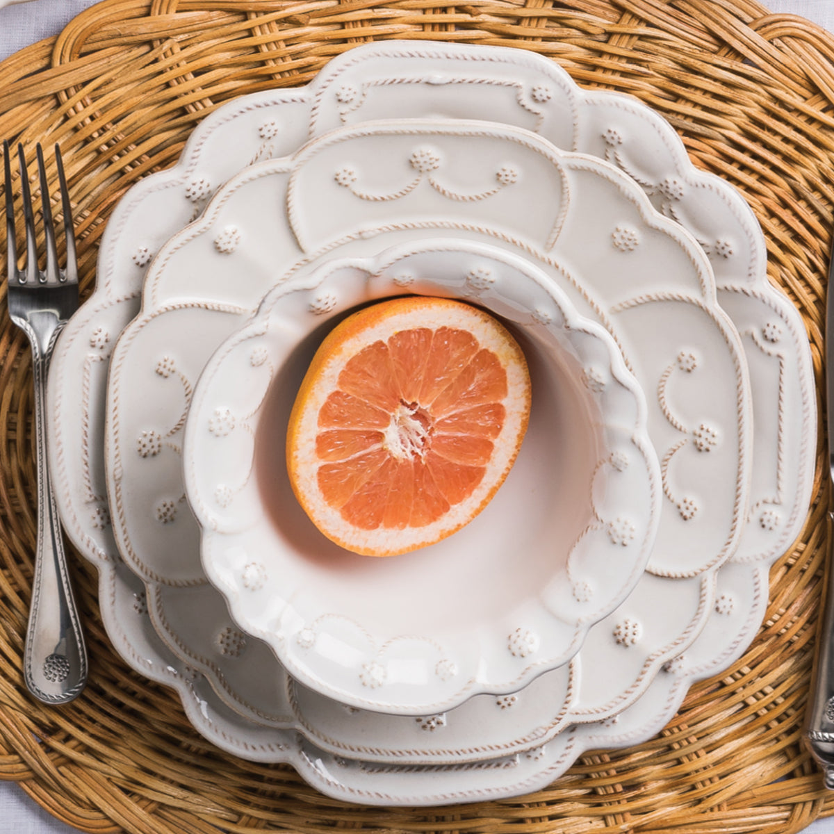 A stacked dinner plate, salad plate and berry bowl, show off the lavish edge detailing of the Jardins du Monde collection. The iconic berries and threads on majestic shapes create the illusion of a blooming flower stacked upon a tan rattan placemat.