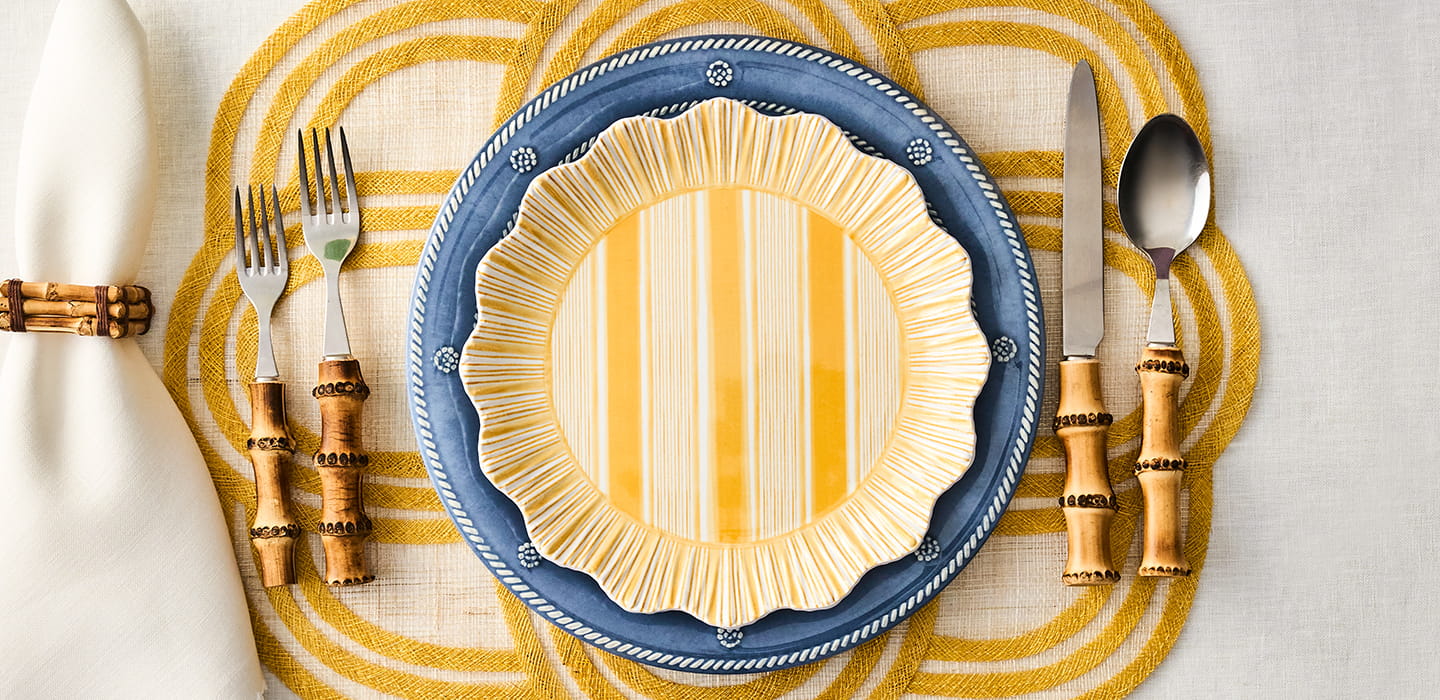 Dining table setting with a blue and yellow plate, cutlery, and decorative elements on a yellow and white striped placemat.