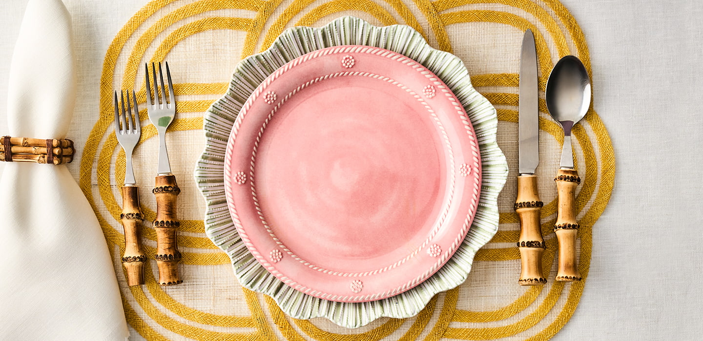 Pink plate with decorative rim on a yellow and white checkered tablecloth, accompanied by cutlery with wooden handles.
