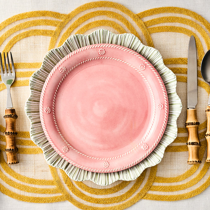 Pink plate with decorative rim on a yellow and white checkered tablecloth, accompanied by cutlery with wooden handles.