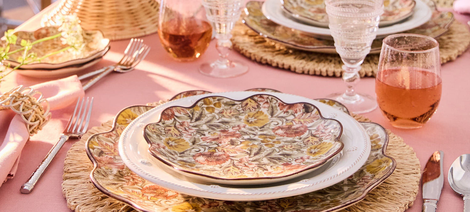 Elegant table setting with floral plates, glasses, and cutlery on a pink tablecloth.