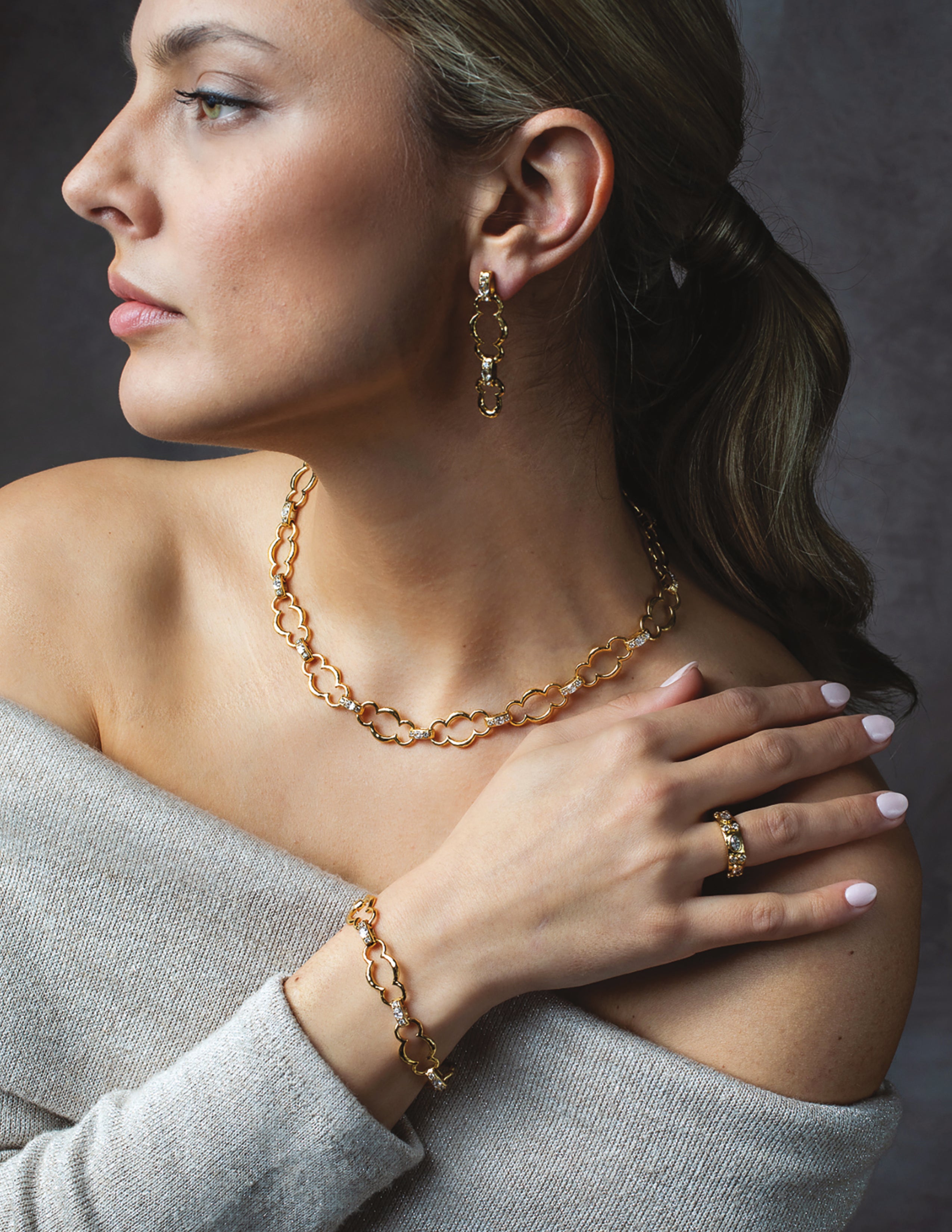 A woman in profile wearing a gold chain necklace, matching gold drop earrings, and a gold bracelet with a ring, styled with a gray off-the-shoulder sweater against a dark gray background.