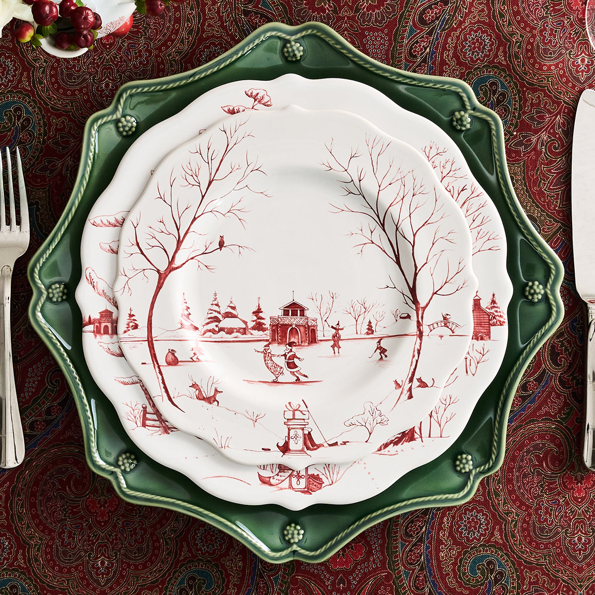 Three stacked ceramic plates with scalloped edges, featuring a red winter landscape toile de Jouy print on the center and middle plates, and a solid dark green plate underneath, set on a red paisley tablecloth.