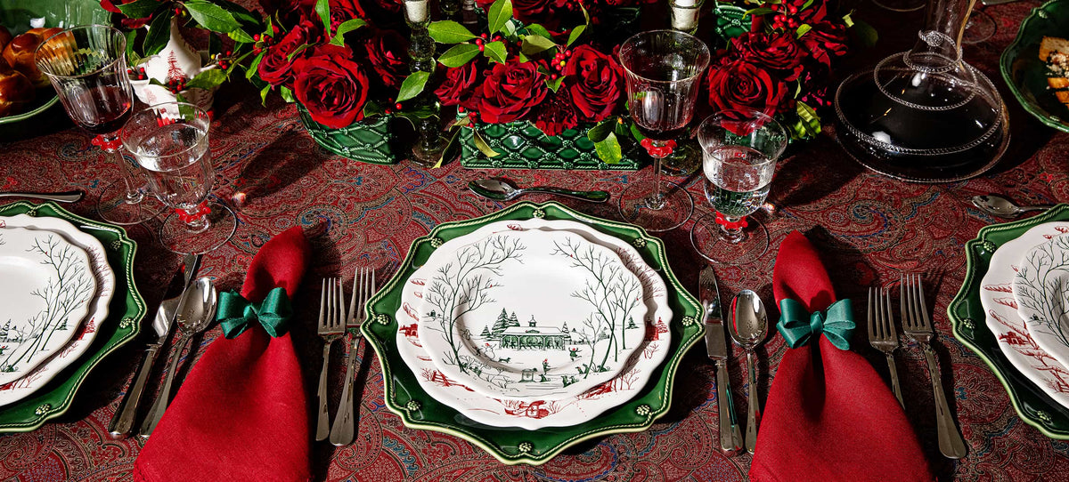 A festive holiday table setting featuring green and white patterned ceramic plates, red napkins tied with green bows, and a red patterned tablecloth decorated with red roses and greenery.