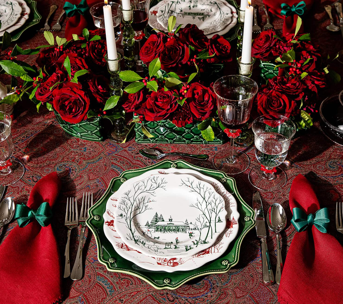 A festive holiday table setting featuring a centerpiece of deep red roses and greenery, a red patterned tablecloth, and a place setting with green and white patterned plates, red napkins tied with teal bows, and silver cutlery.