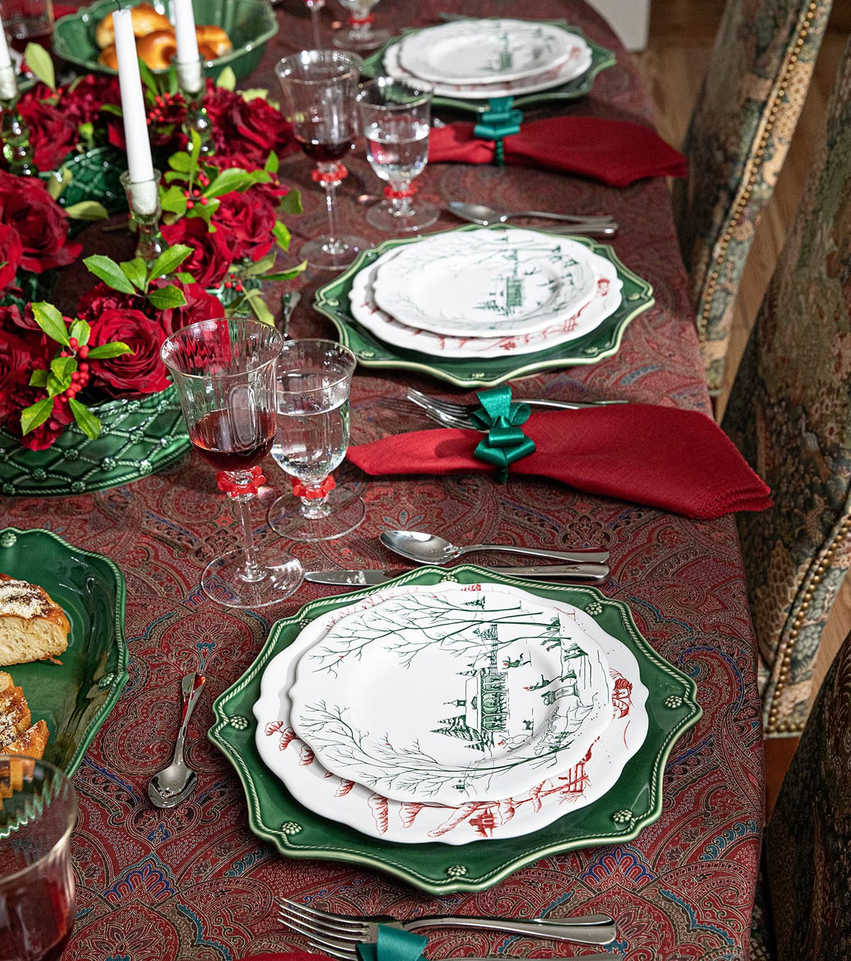 A festive holiday table setting featuring green and white patterned plates, red napkins, green napkin rings, and a red paisley tablecloth decorated with red roses and white candles.