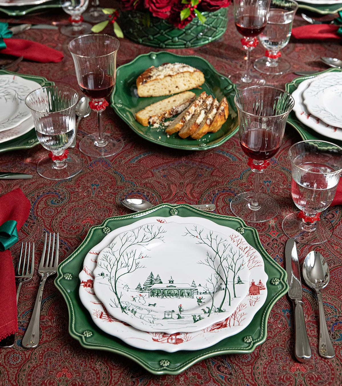 A festive holiday table setting featuring a stack of plates with a green scalloped charger plate, a white plate with a green winter landscape illustration, and silver cutlery on a red patterned tablecloth.