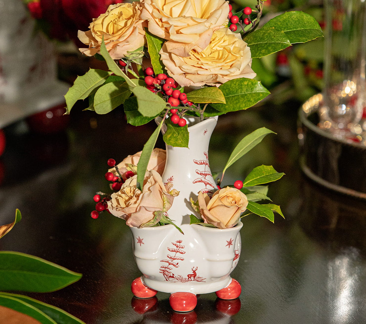 A decorative white ceramic vase with red holiday illustrations, filled with pale yellow roses, green leaves, and red berries, set on a dark reflective tabletop.