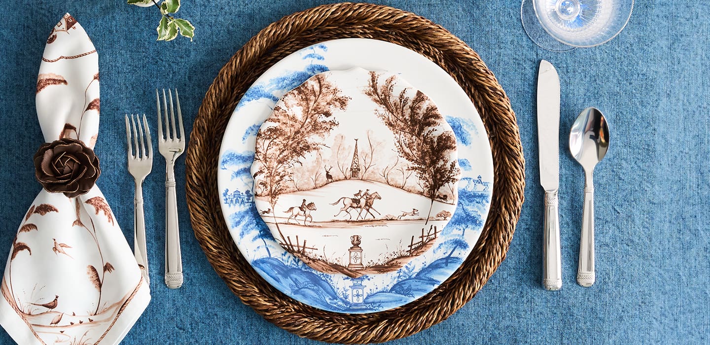 A beautifully styled table setting featuring a round ceramic plate with a brown and blue landscape illustration, layered under a braided brown placemat. The setting includes a patterned napkin with a brown rose, silver flatware, and a textured blue tablecloth.