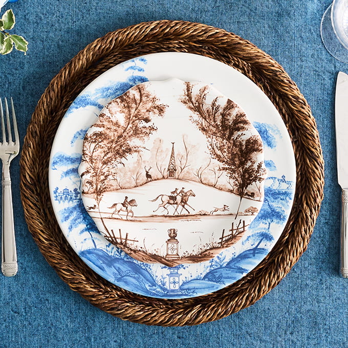 A decorative ceramic plate with a blue and brown landscape print, layered on top of a woven brown placemat over a blue tablecloth, with silverware partially visible at the edges.