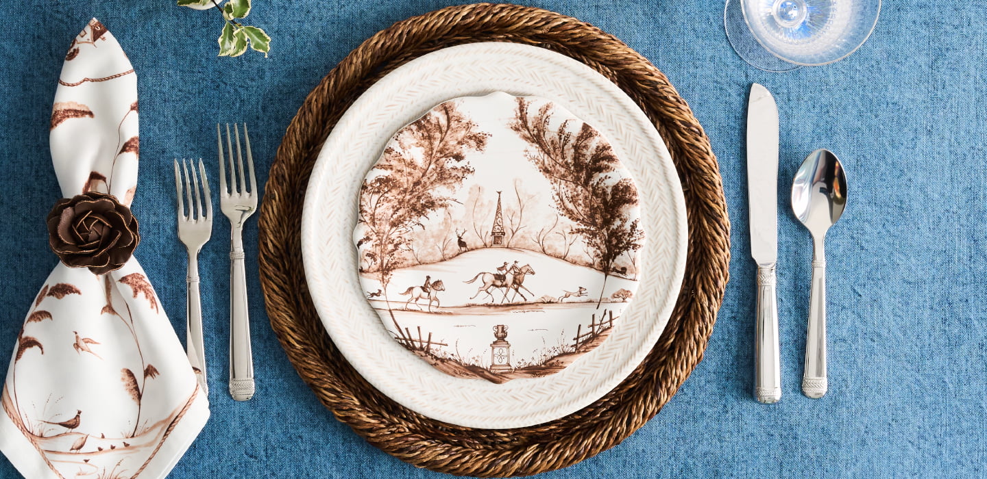 A place setting featuring a white ceramic dinner plate with a brown sepia landscape illustration, layered over a braided brown charger plate. The setting is styled on a textured blue tablecloth with silver flatware (fork, knife, spoon) and a white napkin with a brown floral pattern and a dark brown rose accent.