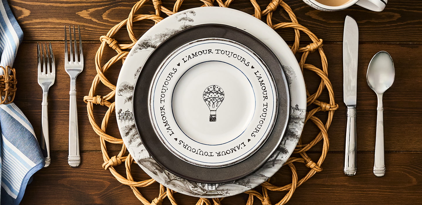 Dining table setting with a decorative plate, cutlery, and woven placemat on a wooden surface.