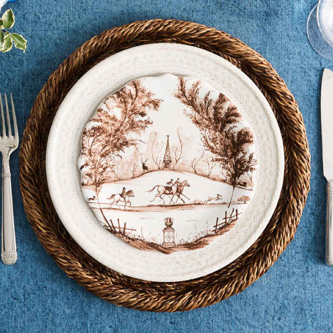 A white ceramic dinner plate with a brown sepia landscape print, layered on a brown woven placemat over a blue tablecloth, with silverware partially visible at the edges.