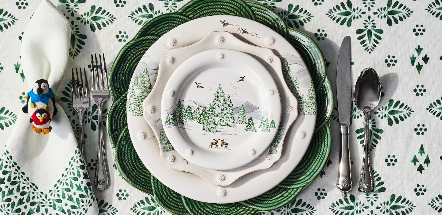 Decorative table setting with patterned Christmas plates, cutlery, and a napkin on a green and white tablecloth.