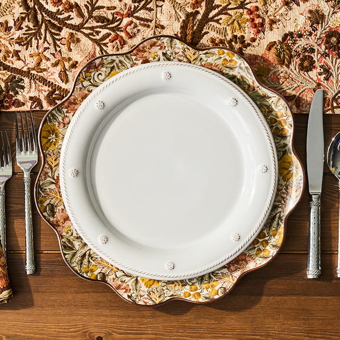 Decorative plate with floral rim on a wooden table with cutlery and a patterned tablecloth.