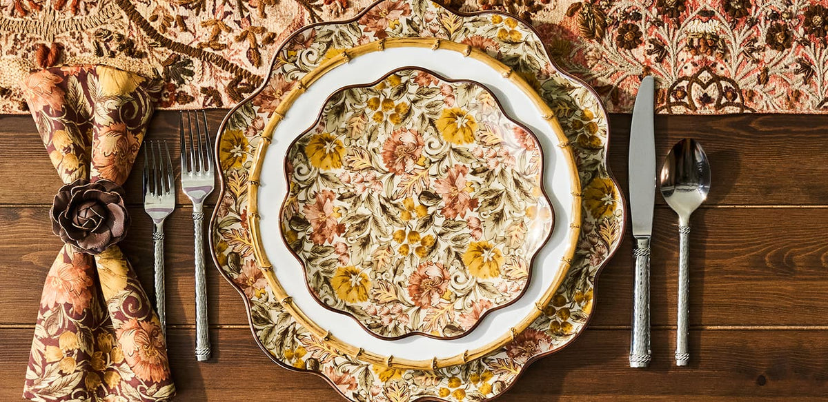 An overhead styled table setting featuring a decorative floral ceramic dinner plate on a larger matching charger plate, accompanied by a patterned napkin tied with a rose-shaped napkin ring, silver flatware, and a rustic dark wood tabletop.