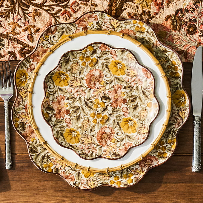 Three scalloped ceramic plates with an intricate floral pattern in yellow, orange, and brown tones, arranged on a wooden table with silver cutlery.