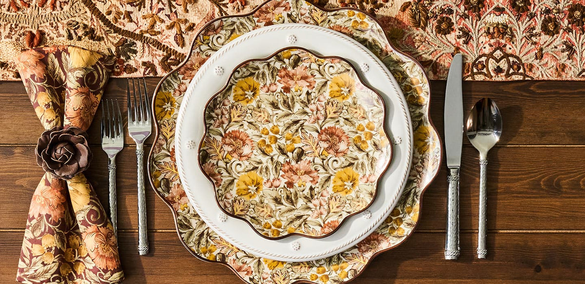 An overhead flat-lay of a decorative place setting on a dark wood table, featuring a scalloped white plate with a floral pattern over matching patterned charger plates, a folded napkin with a rose-shaped ring, silver cutlery, and a patterned textile at the top.