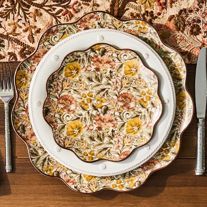 Three stacked decorative ceramic plates with a floral and botanical pattern in yellow, orange, pink, and green, arranged on a wooden table with a silver fork on the left and a knife on the right.