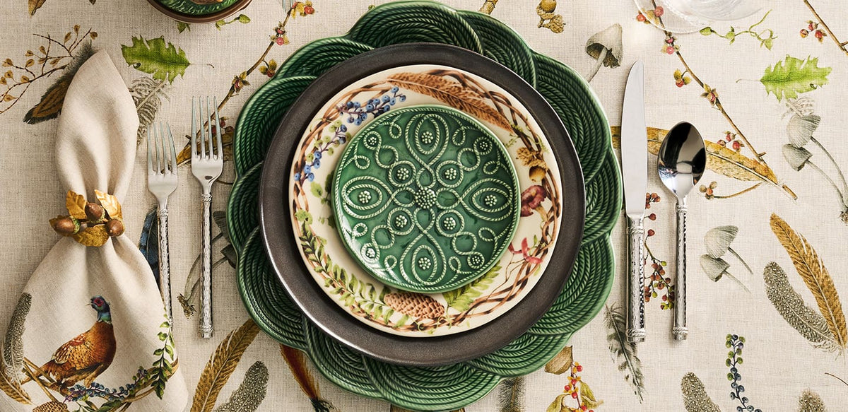 A styled tabletop setting featuring a green ceramic plate with a raised floral medallion pattern, layered over a cream and green patterned dinner plate, a dark green charger plate, a green woven placemat, a napkin with a bird illustration, and silver flatware on a beige botanical tablecloth.