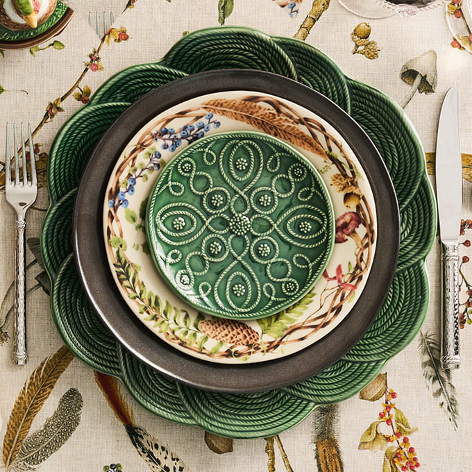 A styled flat-lay of a place setting featuring a green rope-textured charger plate, a dark gray plate, a cream plate with botanical illustrations, and a green patterned plate in the center, surrounded by silver cutlery on a floral tablecloth.