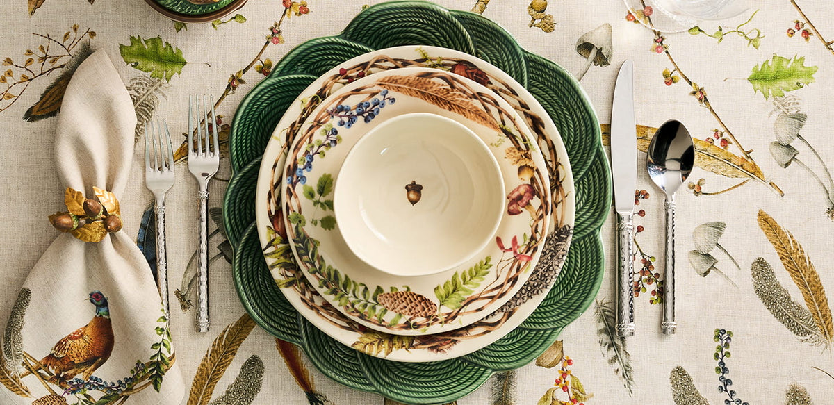 A styled tabletop setting featuring a ceramic dinnerware stack with a cream bowl and plates decorated with woodland botanical and bird motifs, surrounded by green woven placemats, silver flatware, and a patterned tablecloth.