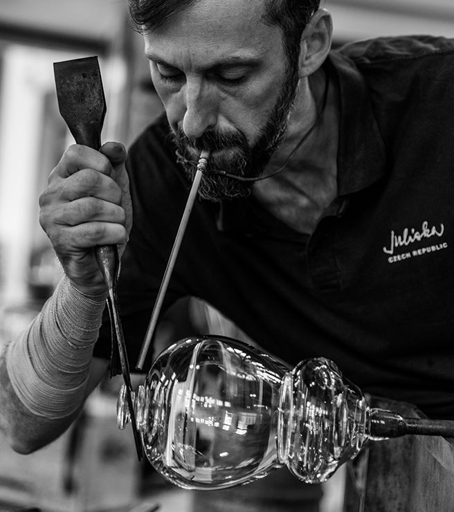 Glassblower shaping a glass object with tools, wearing a Juliska shirt.