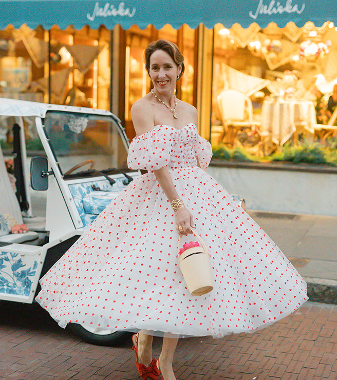 Woman in a white dress with red polka dots standing in front of a vintage car and building.