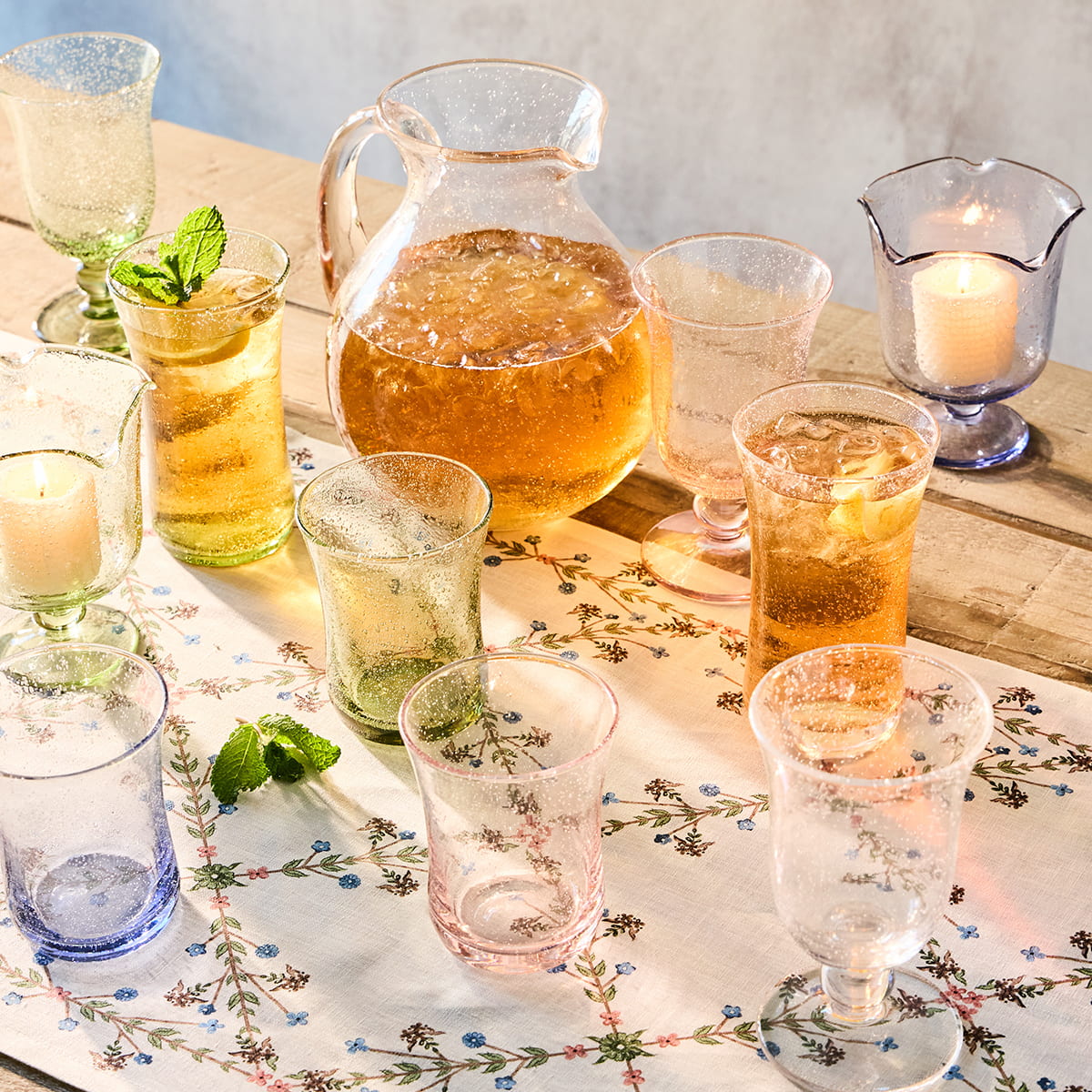 Table setting with glasses, pitcher, and candles in clear, blue and green on a floral tablecloth