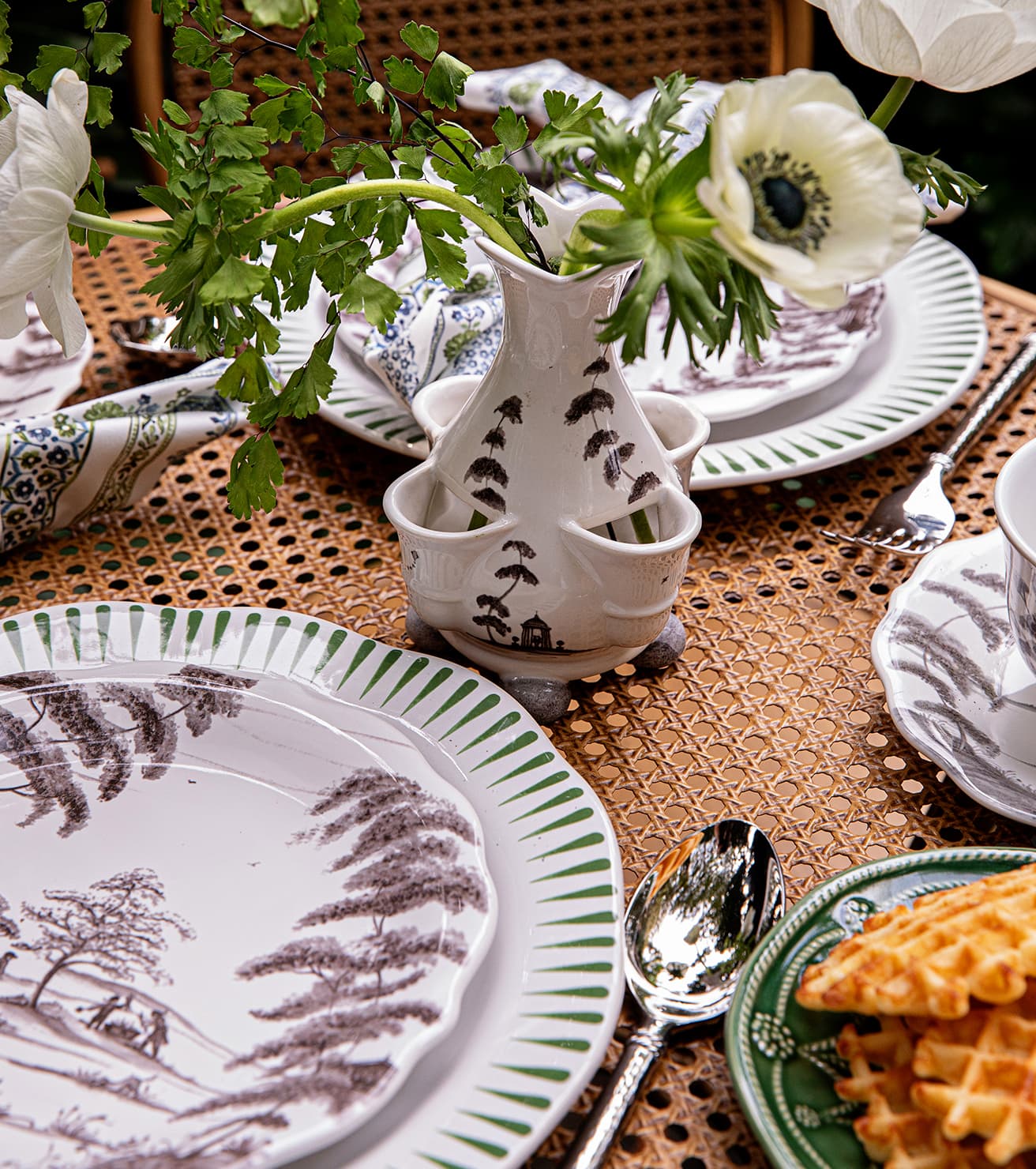 Table setting with decorative plates, a vase with flowers, and waffles on a woven tablecloth.