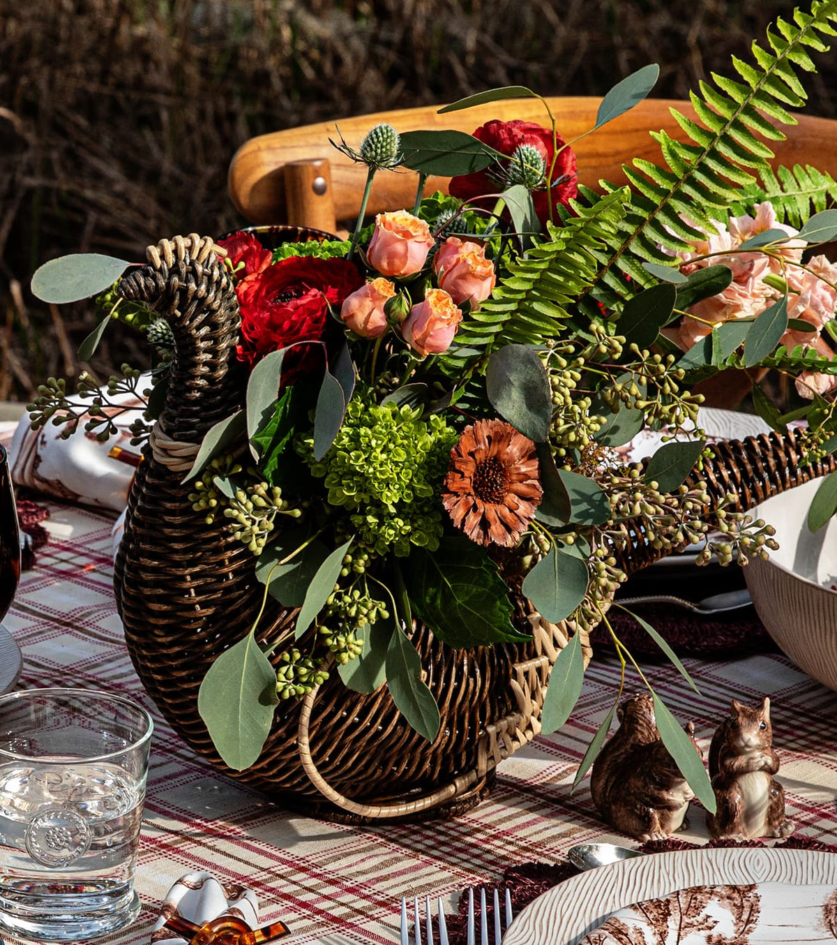 Wicker basket shaped like a bird filled with assorted flowers and greenery, placed on a checked tablecloth among dishes and glassware