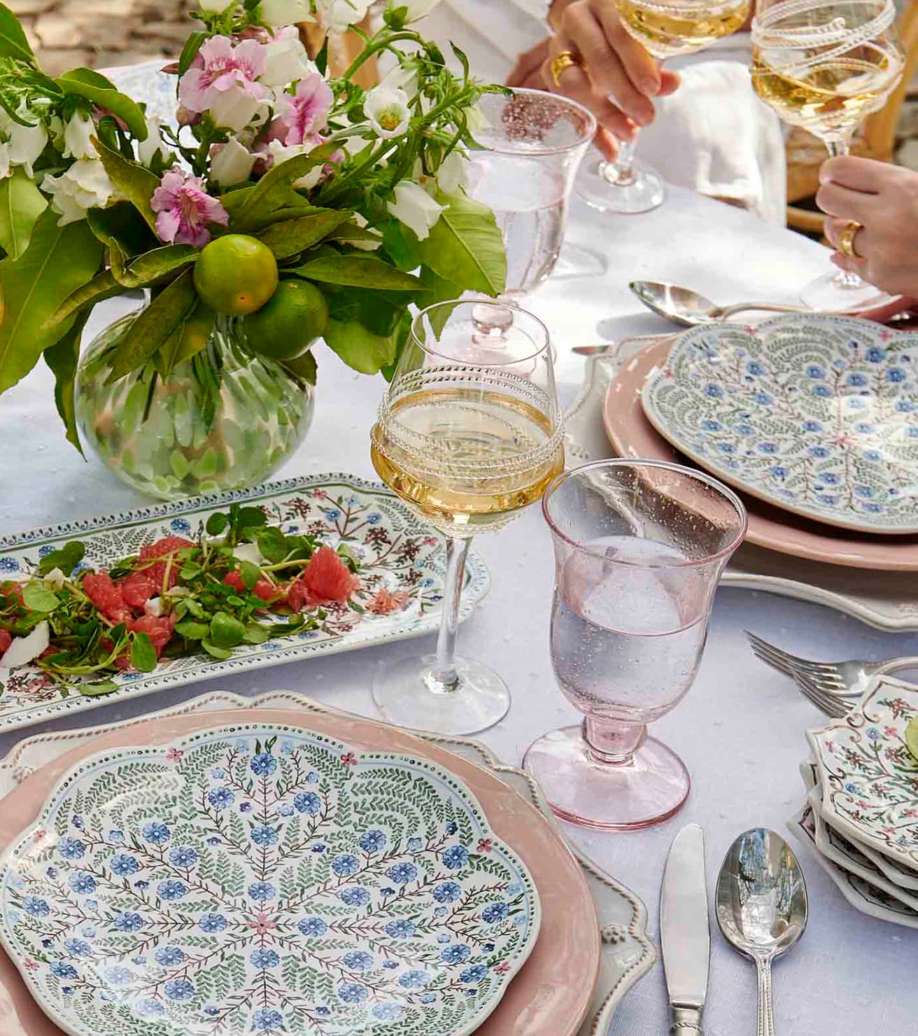 Elegant outdoor dining table setting with floral arrangements, glasses of white wine, and decorative plates.