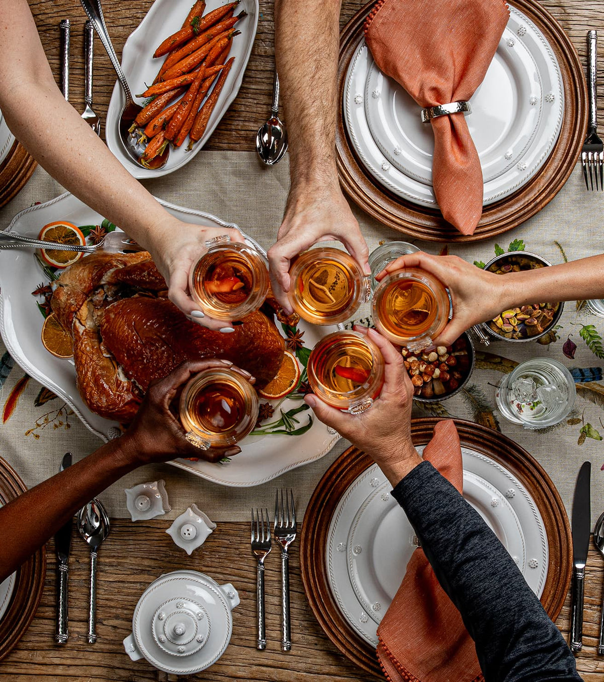 An overhead view of a dining table during a meal, with multiple people's hands holding up glasses of amber-colored beverage to toast. A large platter of roasted turkey is on the left, sliced carrots on a plate at the top left, stacked white plates with orange napkins at the top right, and cutlery and glassware arranged around the table on a rustic wood surface.