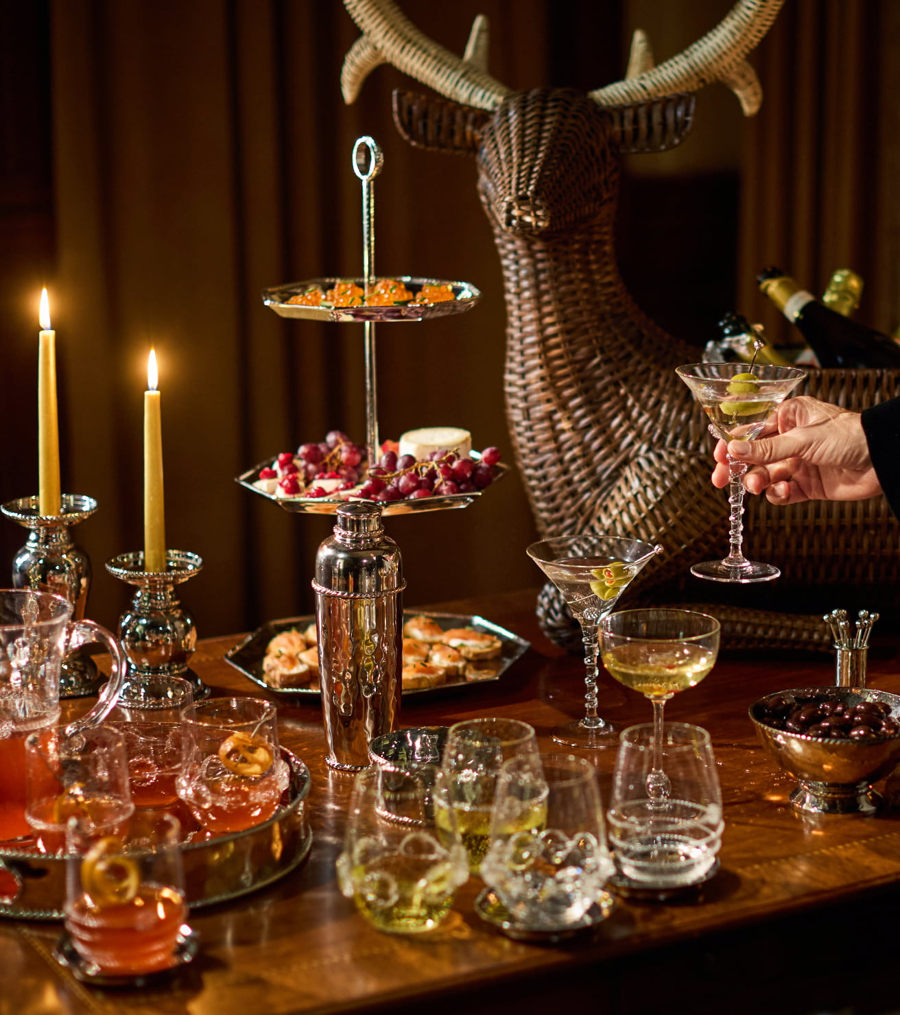Dining table with various drinks and appetizers, lit by candles, with a deer head mount in the background.