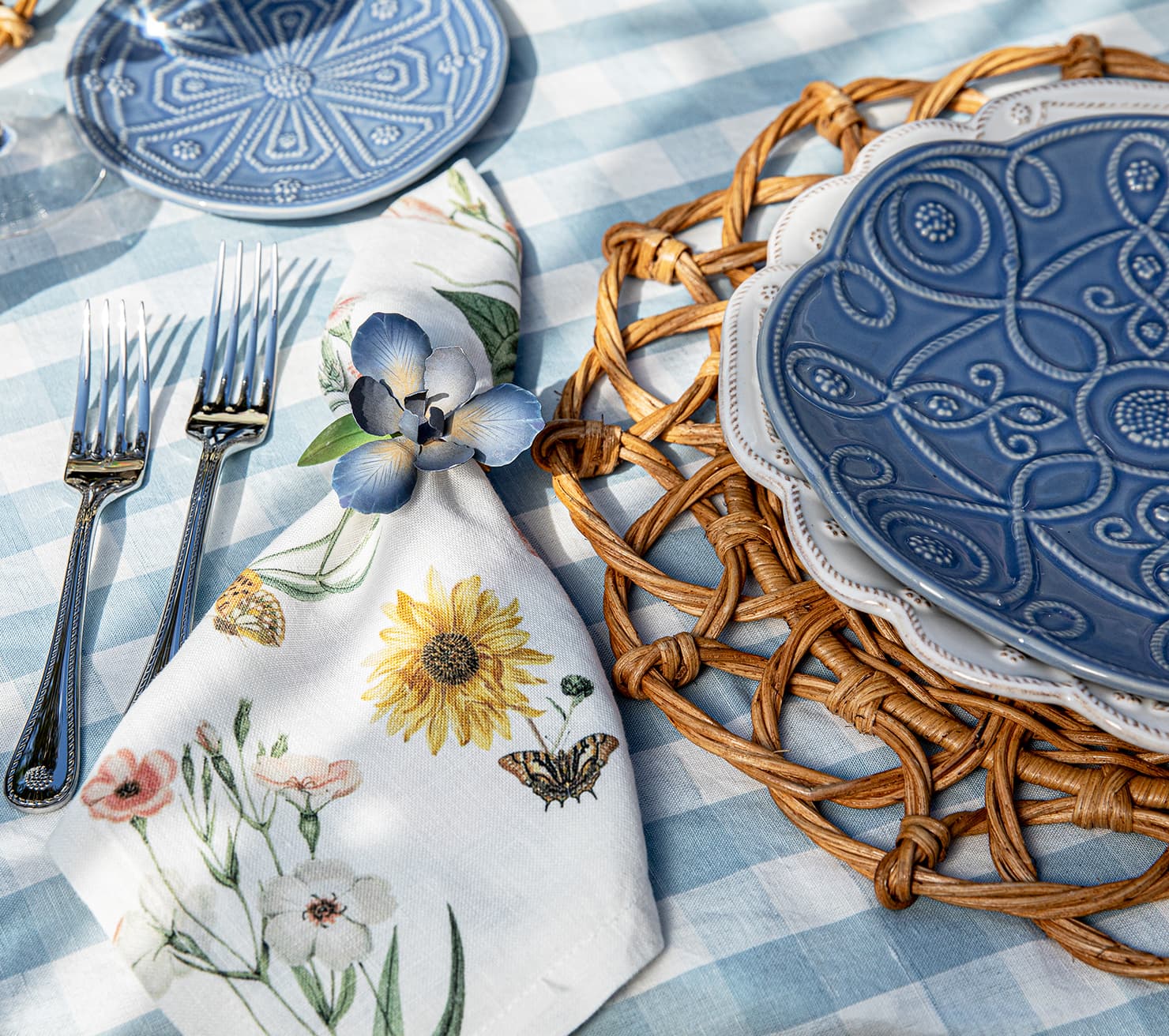 Table setting with floral napkin, blue plates, and wicker placemat on checkered tablecloth