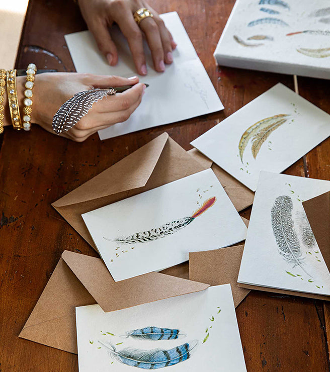 Feather-themed cards and envelopes on a wooden surface with hands interacting.
