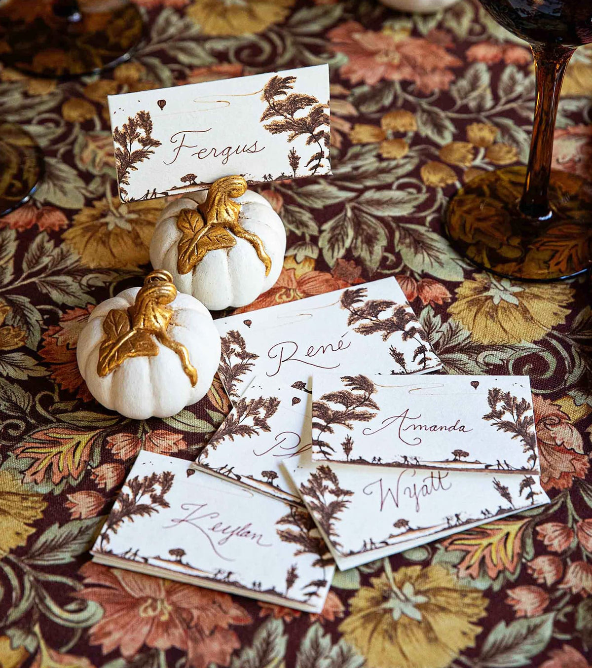 Place cards with names like "Fergus," "René," "Amanda," "Wyatt," and "Keylan" are displayed on a floral tablecloth, held up by small white pumpkins with gold stems and leaves, suggesting an autumn celebration or dinner party.