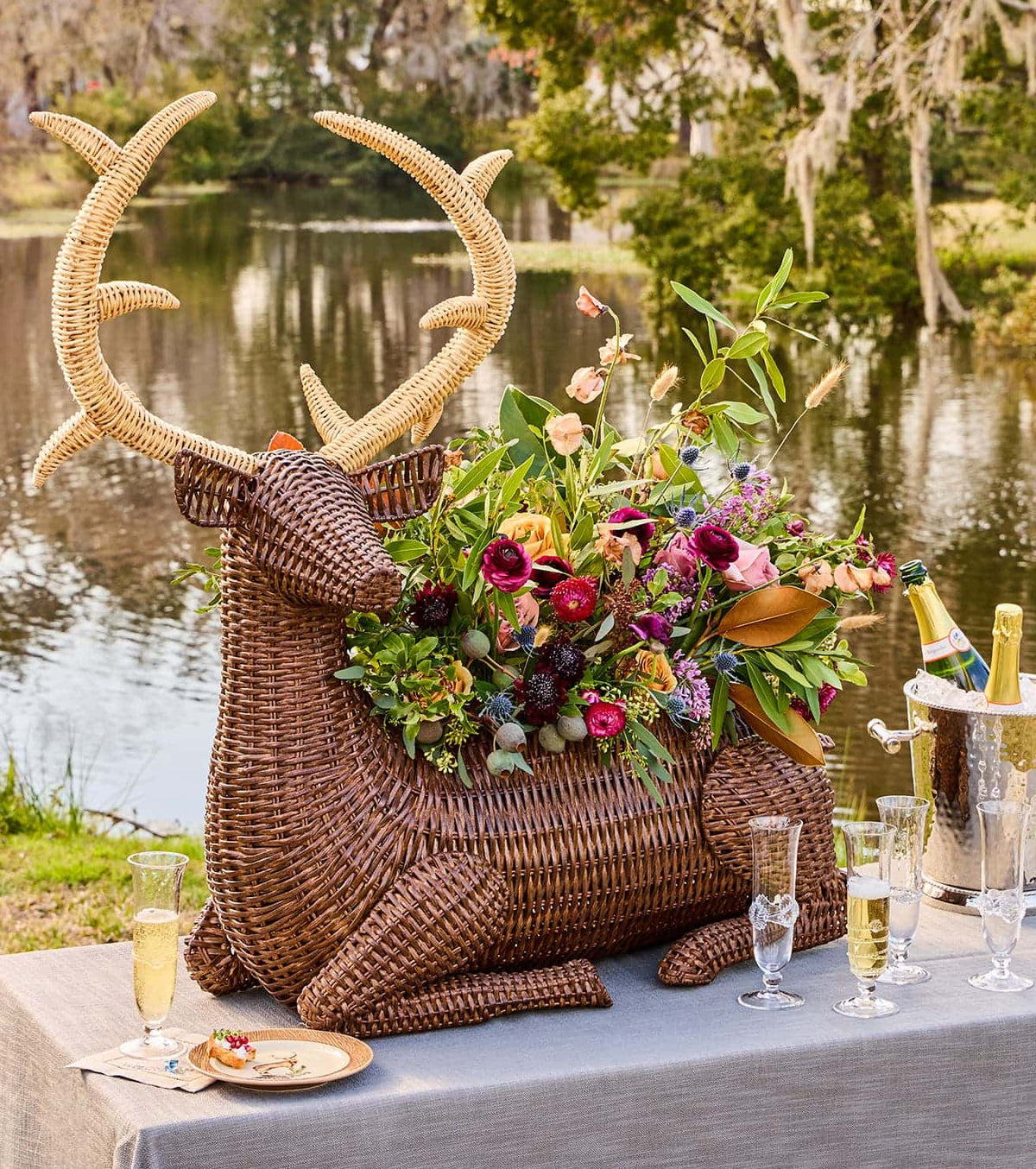 A large woven deer-shaped centerpiece with antlers and a lush floral arrangement sits on a table outdoors by a lake, styled with champagne flutes, a champagne bucket, and a small plate of food.