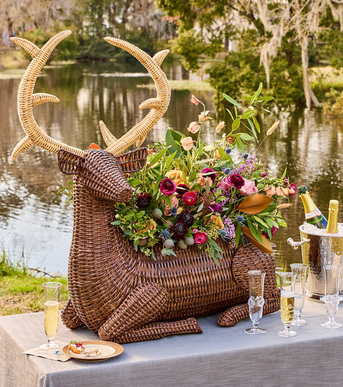 A large woven deer-shaped centerpiece with antlers and a lush floral arrangement sits on a table outdoors by a lake, styled with champagne flutes, a champagne bucket, and a small plate of food.