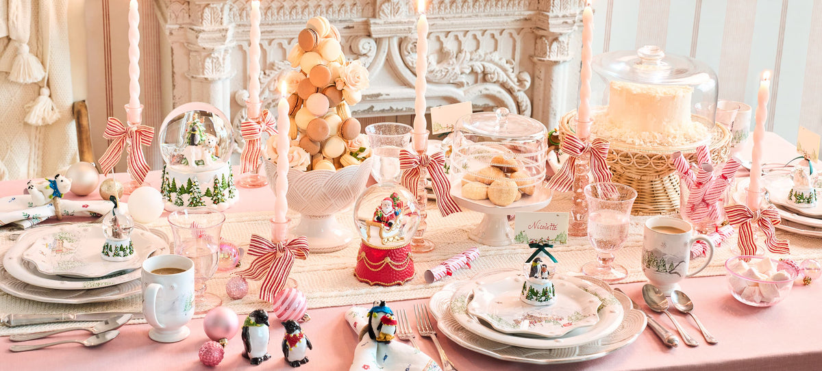 A beautifully styled pastel pink holiday table setting featuring white plates, pink and white striped ribbons, penguin figurines, a snow globe, and a tower of macarons in the background.