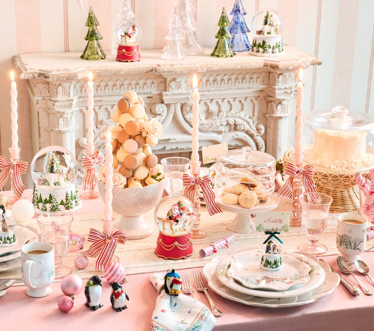 A beautifully styled holiday table setting with a pink tablecloth, white plates, teacups, macarons, snow globes, and miniature Christmas trees, arranged in front of an ornate white fireplace.