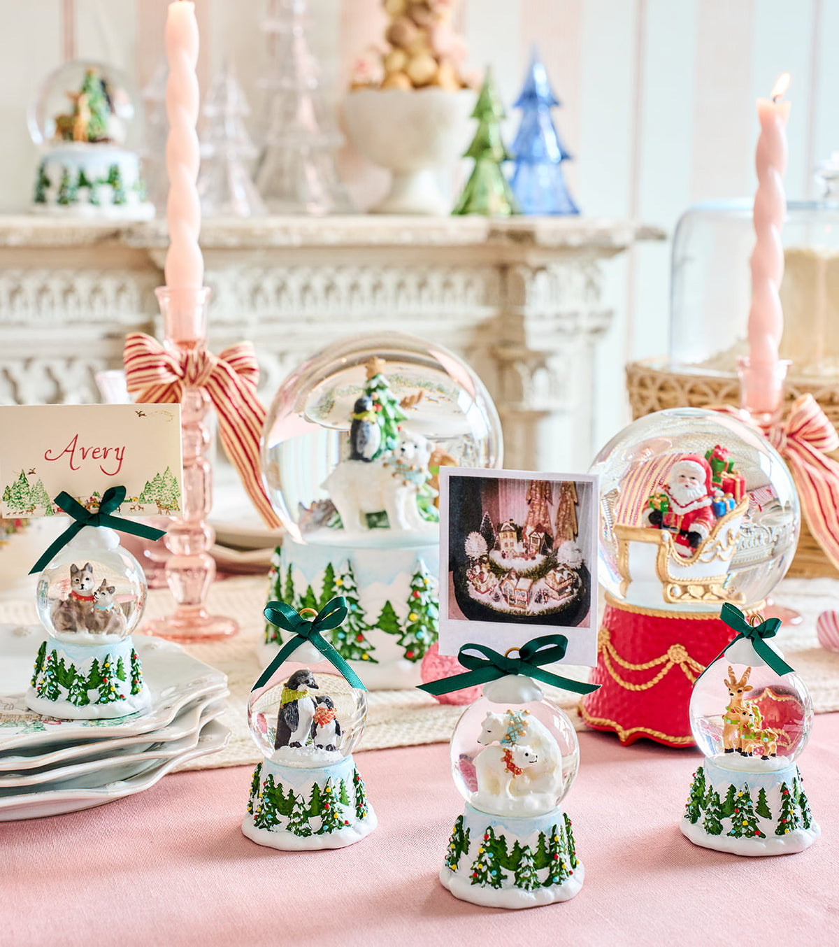 A styled holiday tabletop display featuring multiple decorative snow globes with festive scenes inside, including Santa in a sleigh, a penguin couple, a snowman and angel, and a reindeer. The globes are arranged on a pink tablecloth with pink candles, ribbons, plates, and a card in a softly lit interior setting.