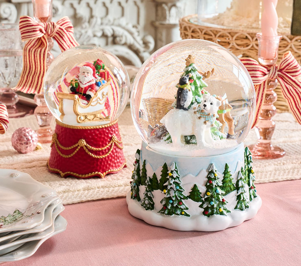 Two decorative Christmas snow globes displayed on a pink table runner. The larger foreground globe has a white base with green pine trees and a white horse-drawn sleigh under a green tree.