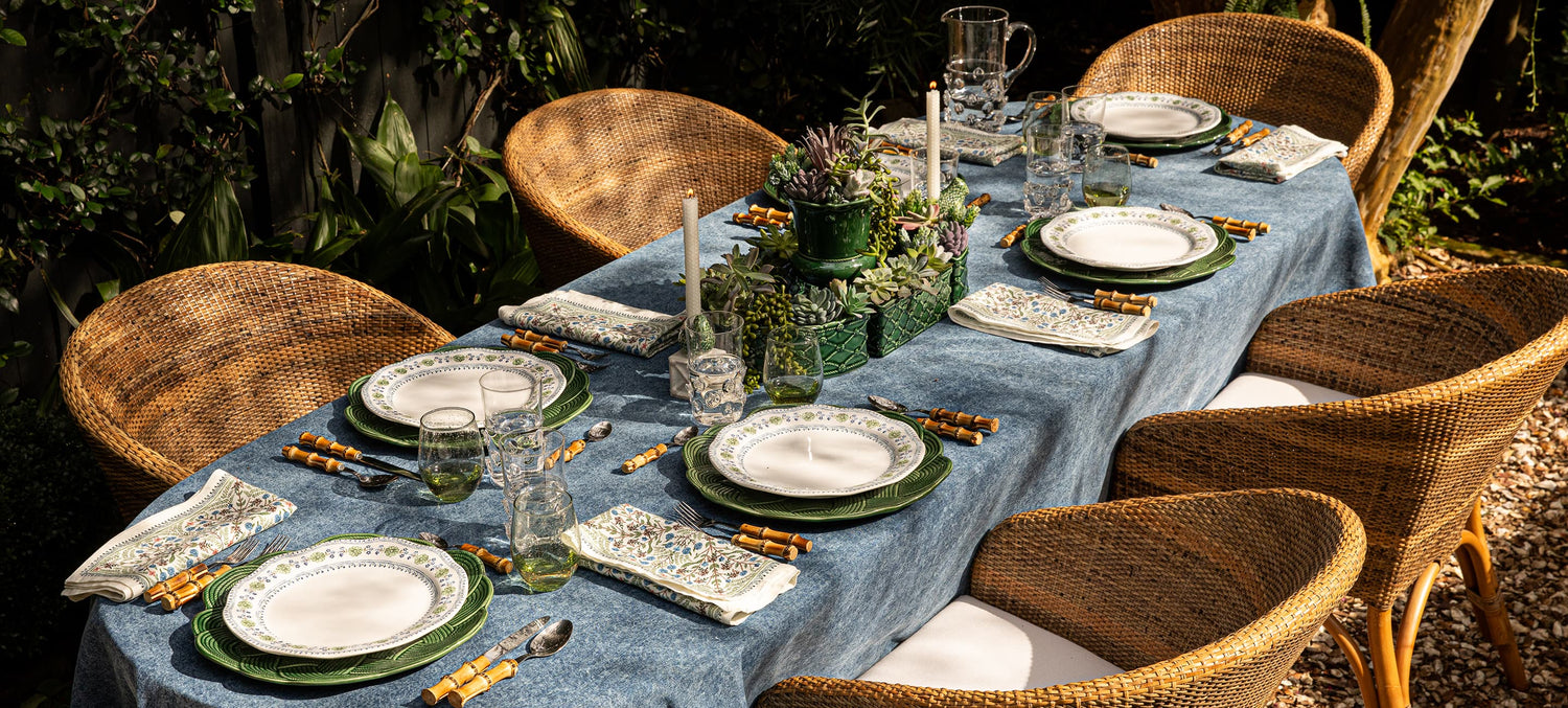 Dining table set for a meal with plates, glasses, and cutlery on a blue tablecloth outdoors.