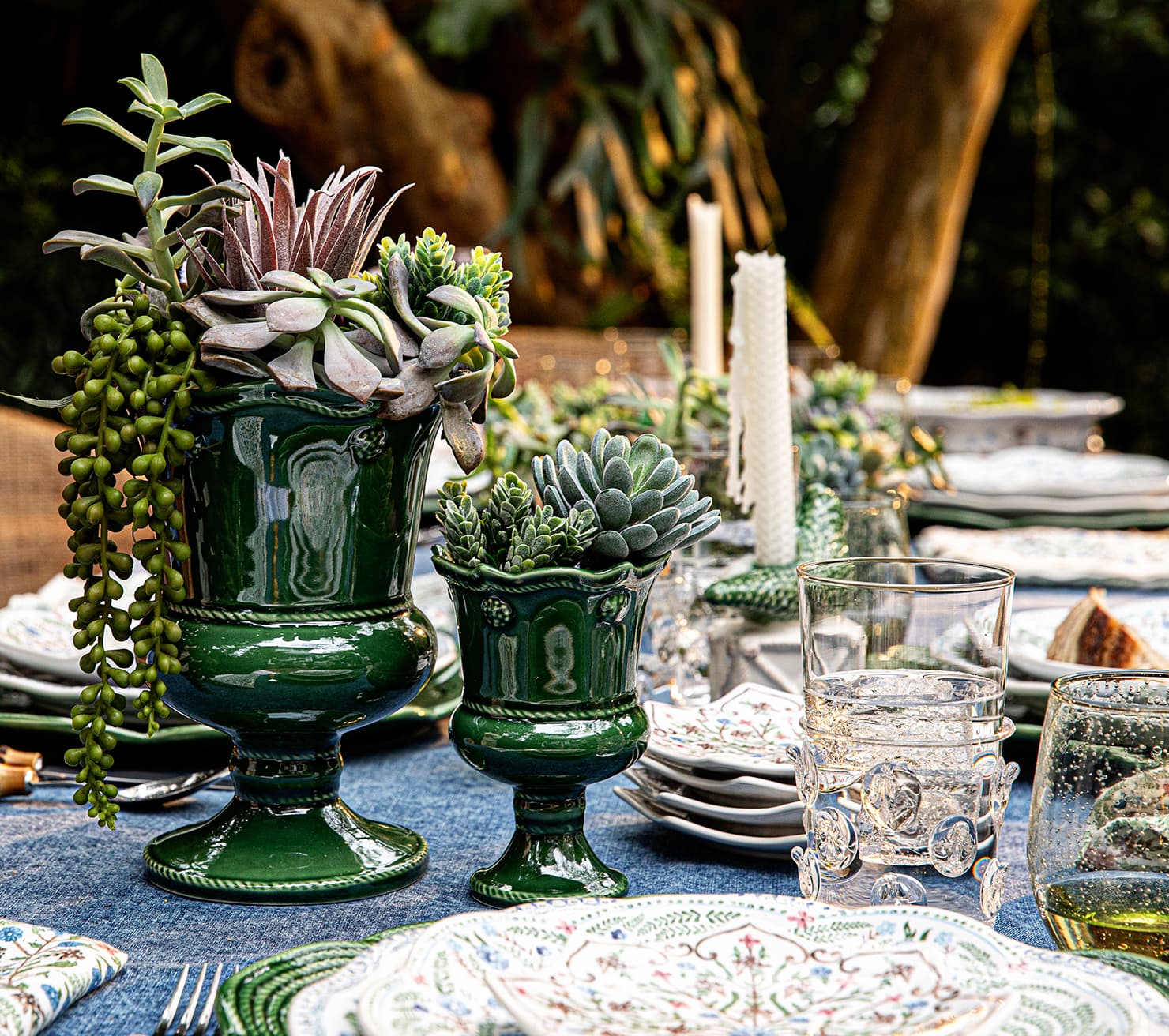 Decorative table setting with green urns, succulents, and glassware on a blue tablecloth.