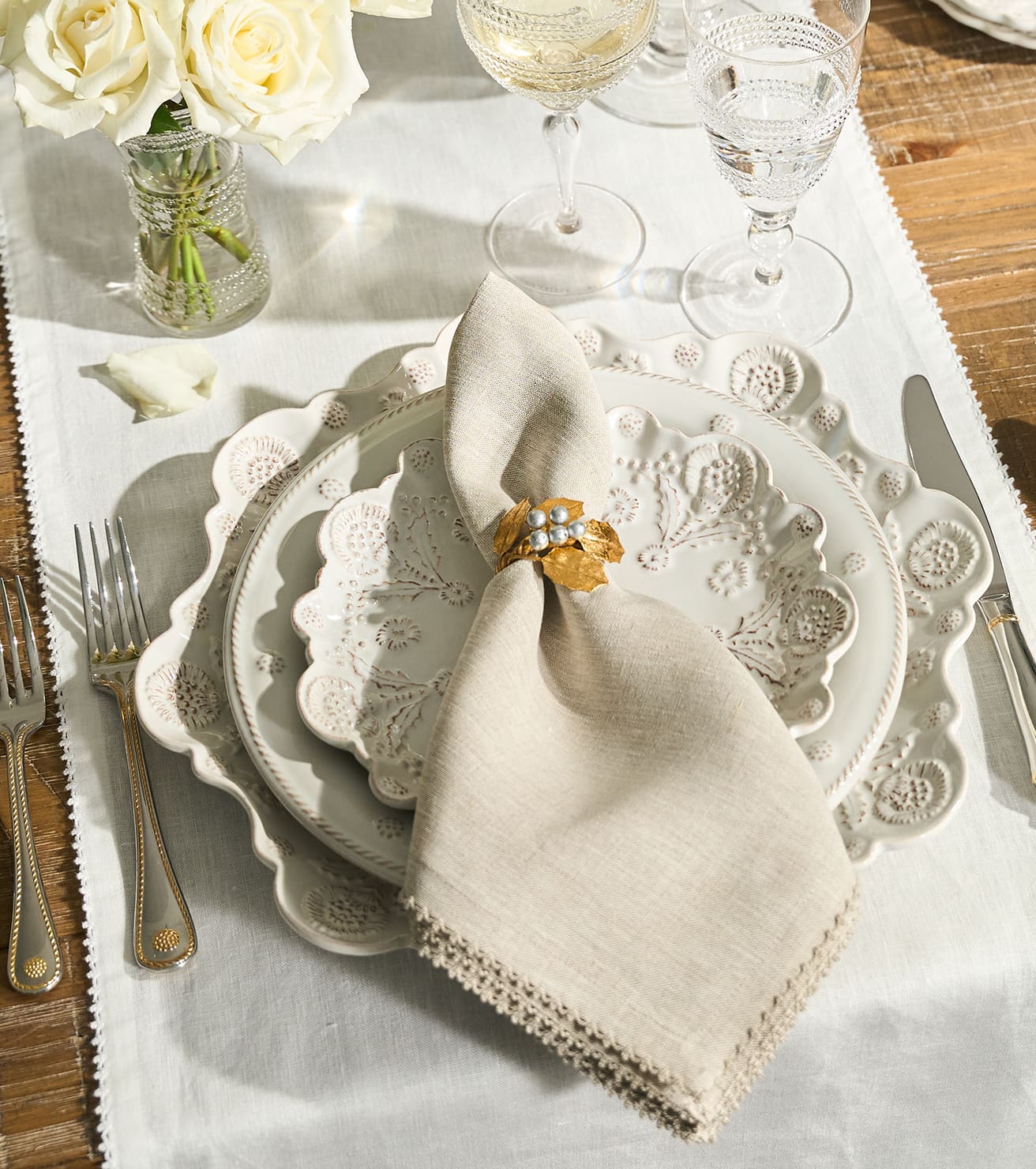 A styled table setting featuring a place setting with a cream linen napkin tied with a gold floral ring, layered on white and cream scalloped ceramic plates, with silver cutlery and a glass vase of white roses on a wooden table.