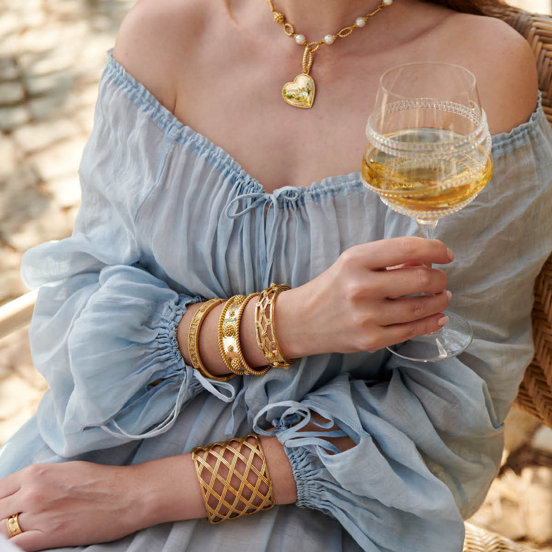 Woman in a light blue dress holding a glass of white wine, wearing gold jewelry.