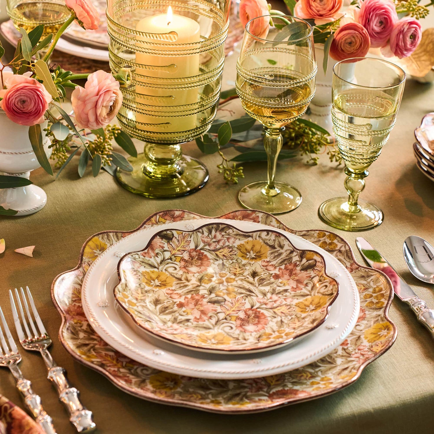 A styled table setting featuring a decorative floral ceramic dinner plate and charger plates with scalloped edges, accompanied by ribbed green glass candle holders, wine glasses, and pink roses on a green tablecloth.