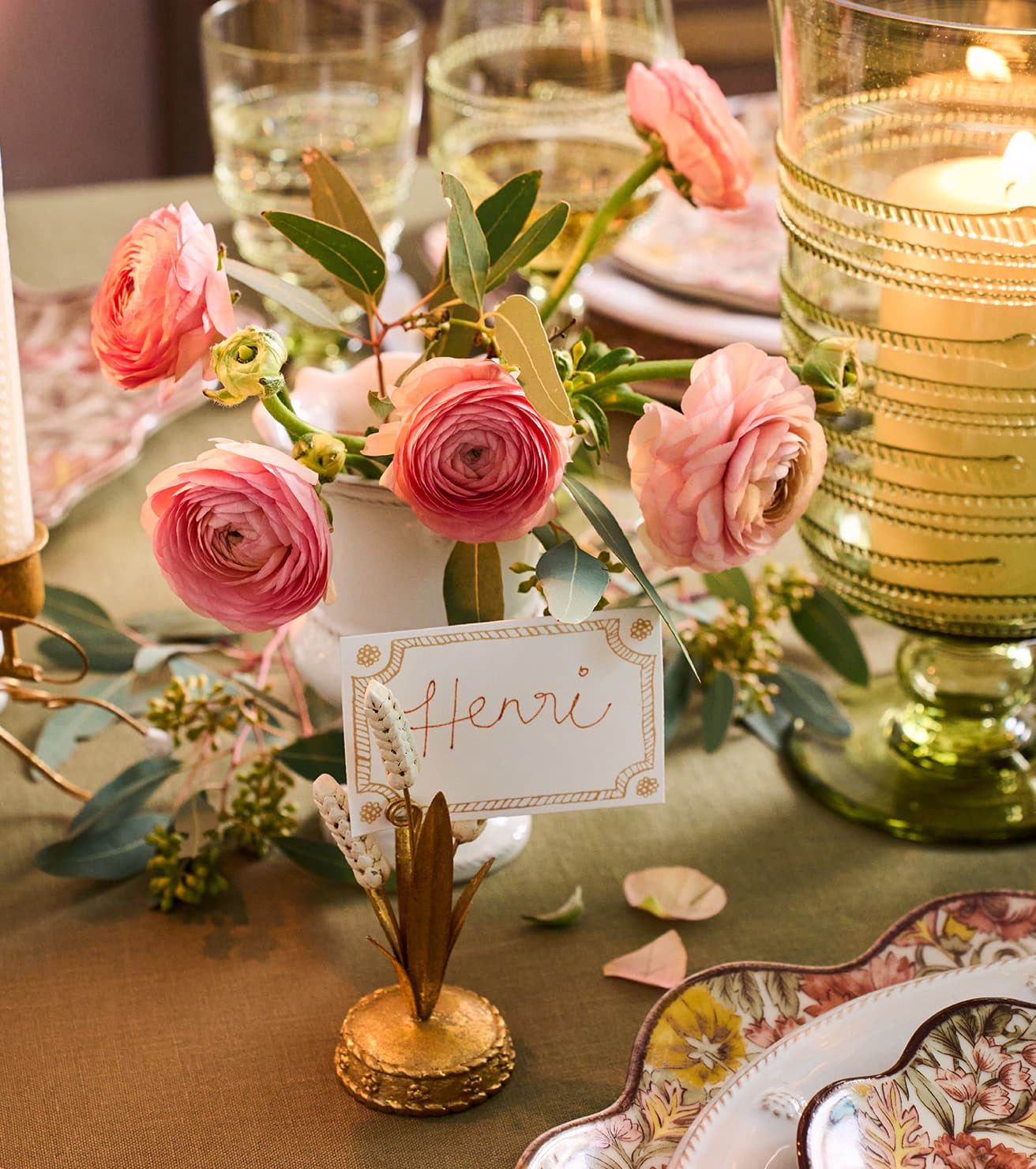A styled table setting with pink ranunculus flowers in a small white vase, a gold candle holder, a green glass candle holder with a lit candle, and a place card reading 'Henri' on a gold leaf stand.