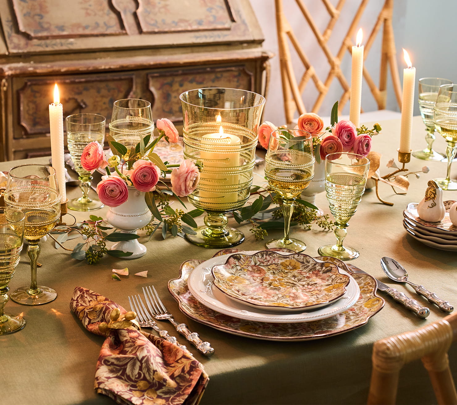 An elegantly styled table setting with vintage-inspired tableware, pink roses, lit candles, and clear glassware in a warm, atmospheric interior scene.